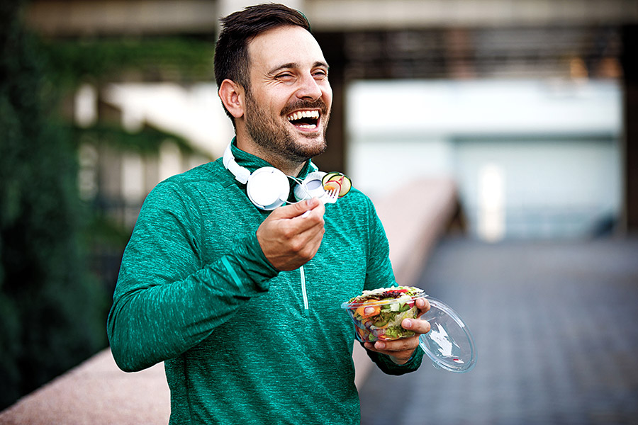 Smiling male in green shirt, white headphones, eating salad.